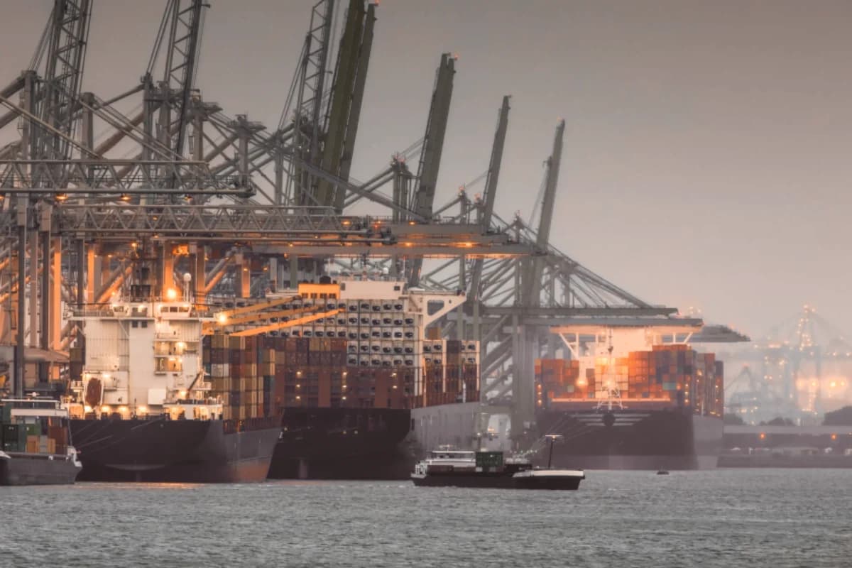 An illuminated container terminal, gantry cranes and a tugboat on the water at dusk.