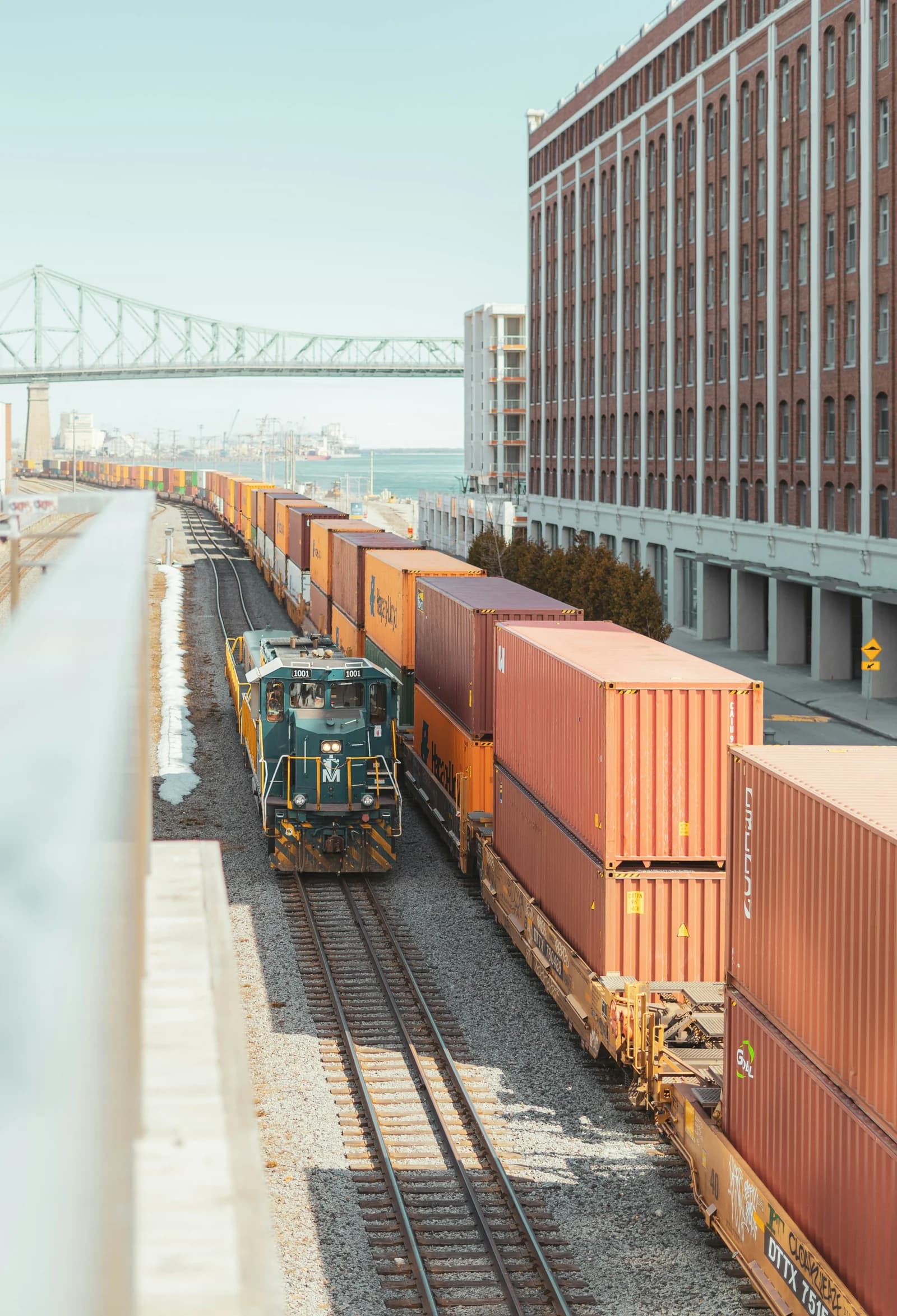 A green locomotive, railway tracks and a long line of wagons carrying orange containers, viewed from above at an angle.